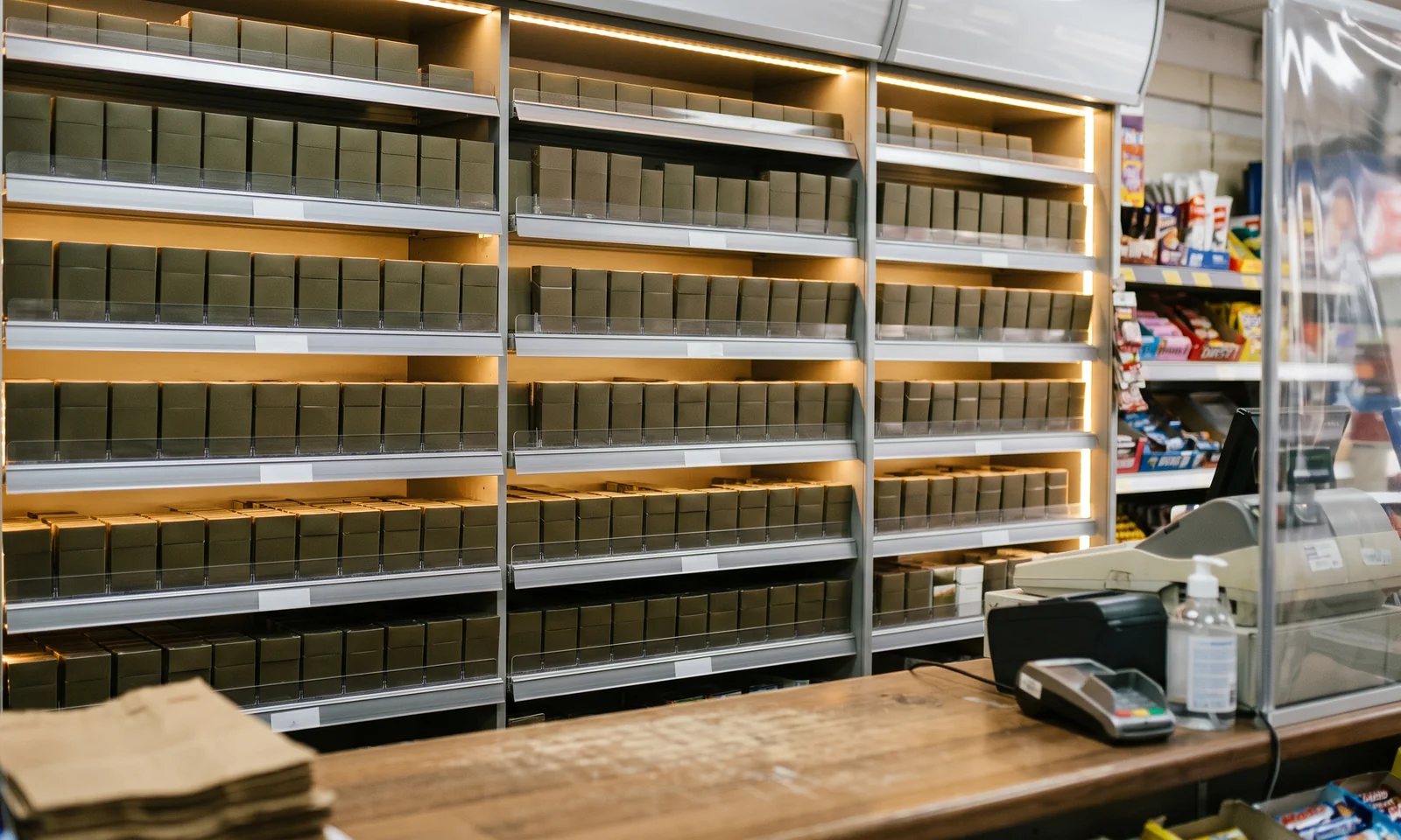A densely stocked tobacco gantry behind the counter of a small convenience shop, rows of plain unbranded packs under warm shelf lighting